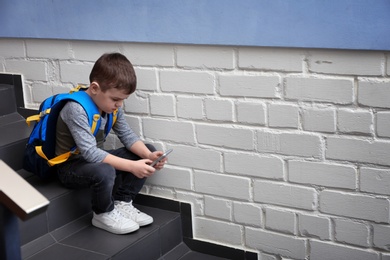 Sad little boy with mobile phone sitting on stairs indoors