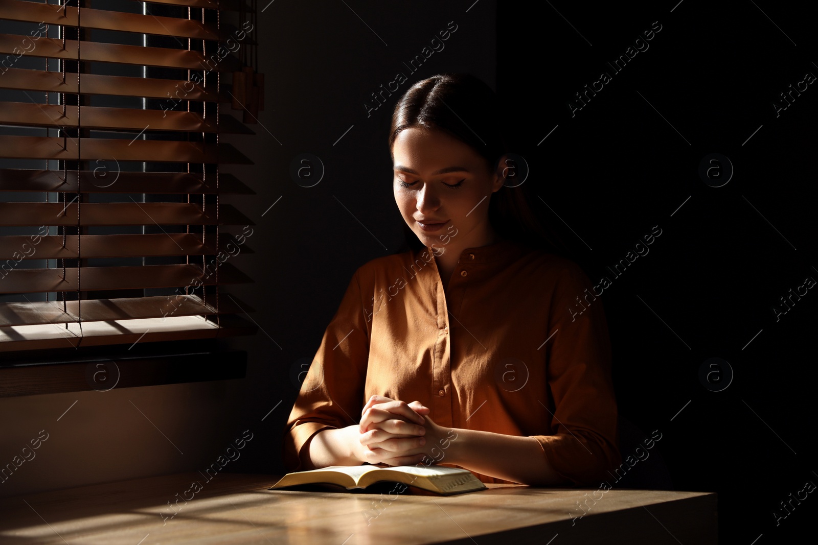 Photo of Religious young woman praying over Bible at wooden table indoors