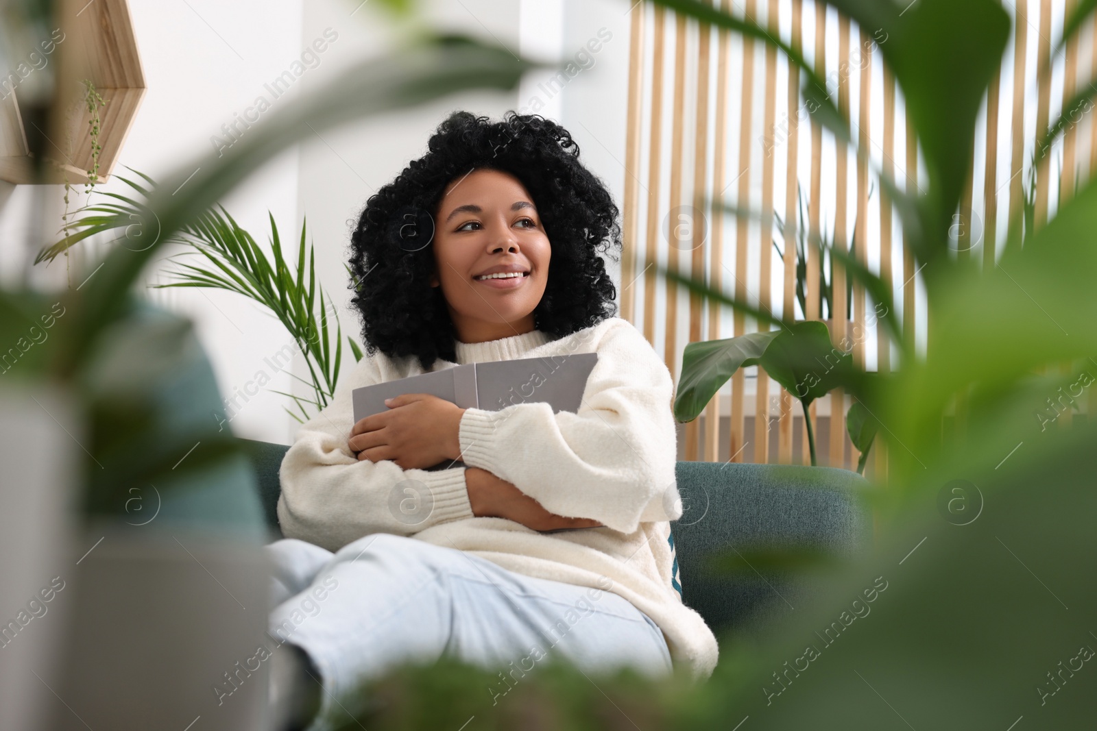 Photo of Relaxing atmosphere. Happy woman with book on sofa surrounded by beautiful houseplants in room