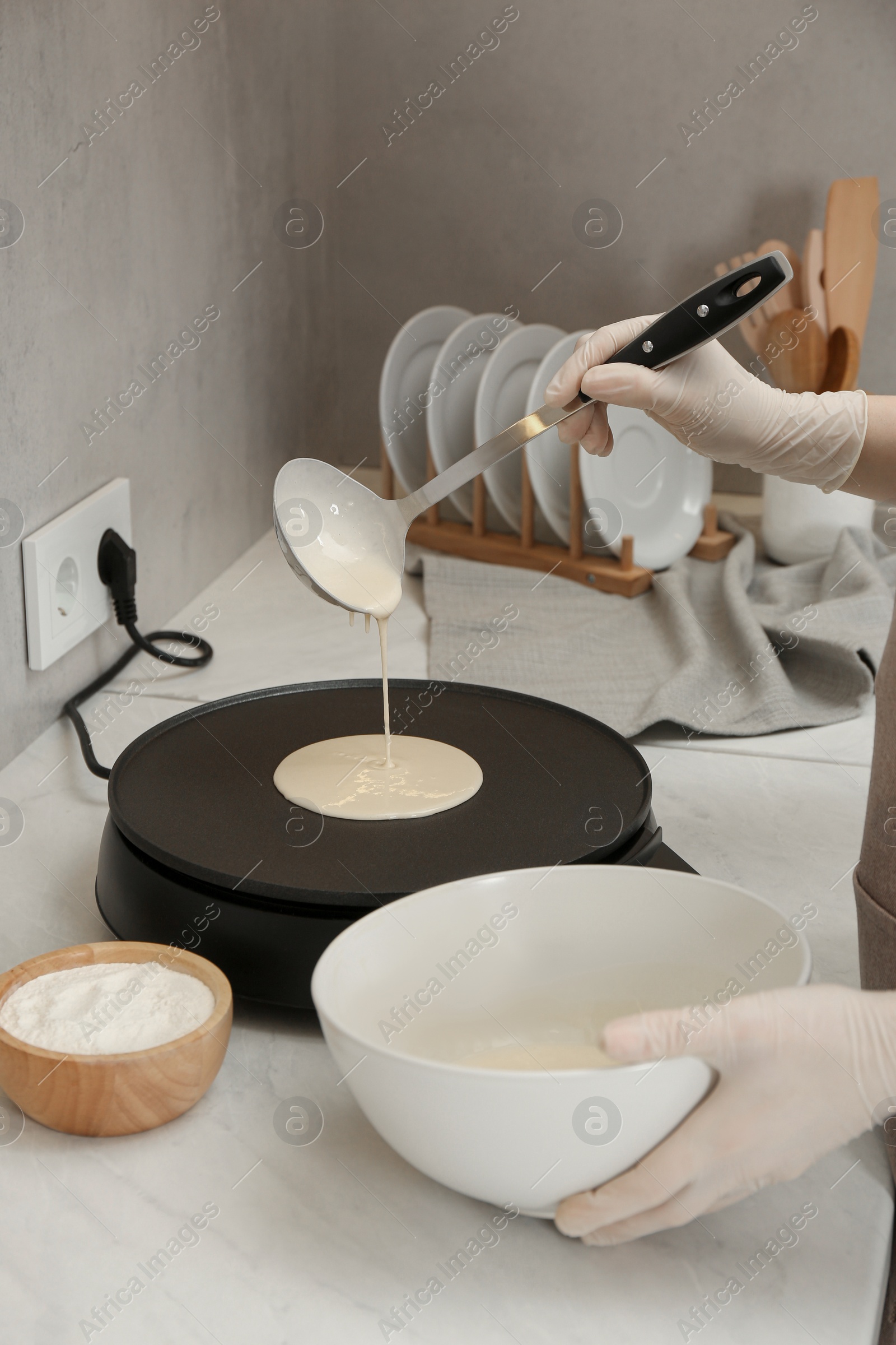 Photo of Woman cooking delicious crepe on electric pancake maker at white marble table in kitchen, closeup