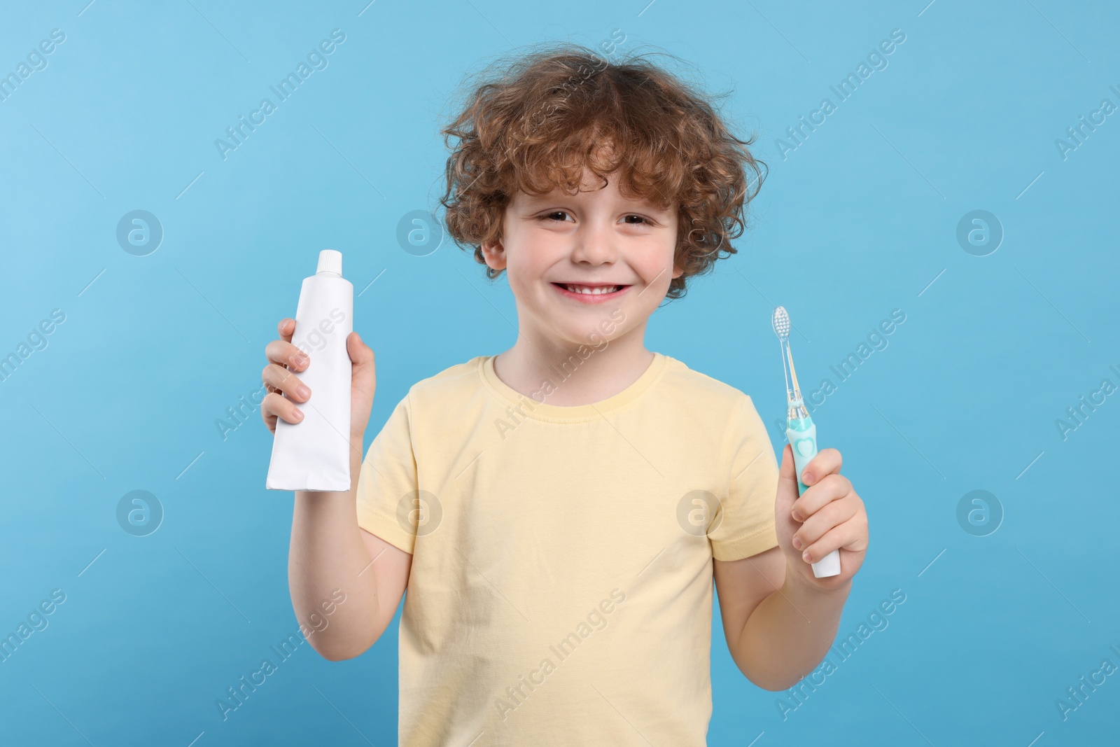 Photo of Cute little boy holding electric toothbrush and tube of toothpaste on light blue background