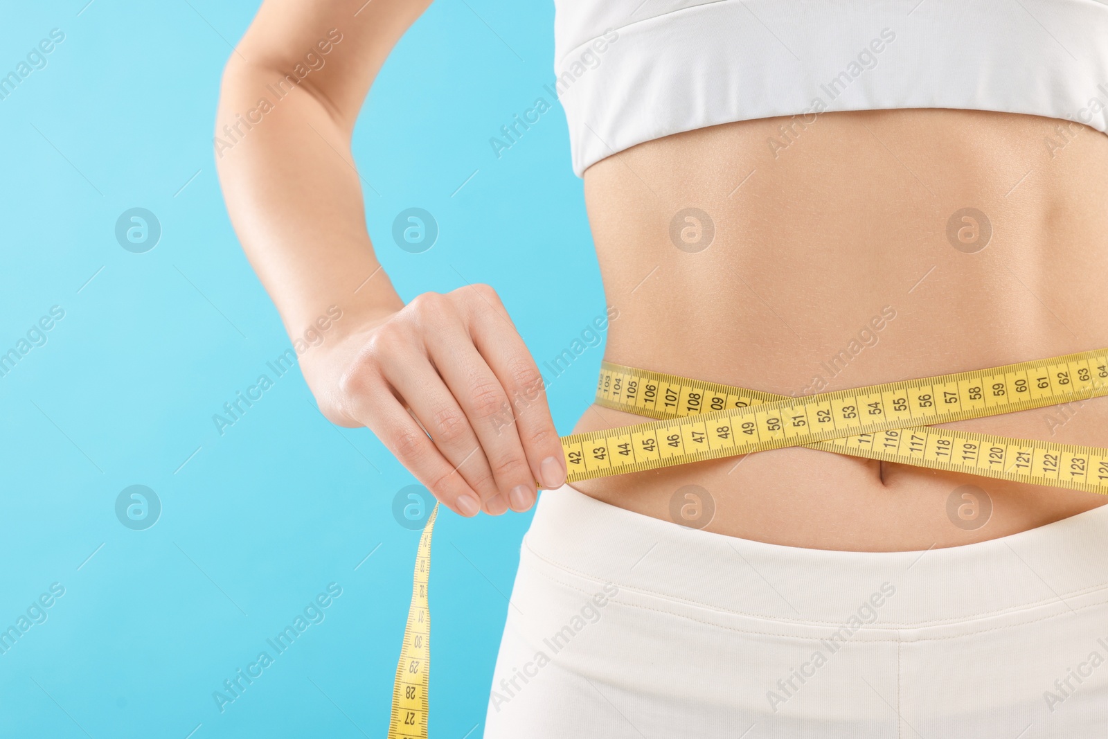 Photo of Slim woman measuring waist with tape on light blue background, closeup. Weight loss