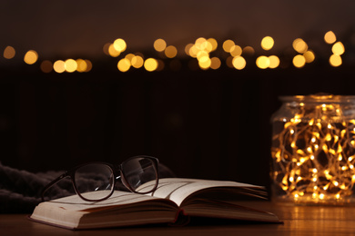 Photo of Eyeglasses and open book on wooden table against festive lights. Space for text