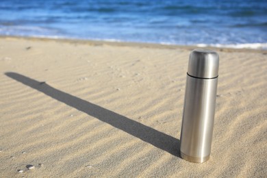 Metallic thermos with hot drink on sand near sea, space for text