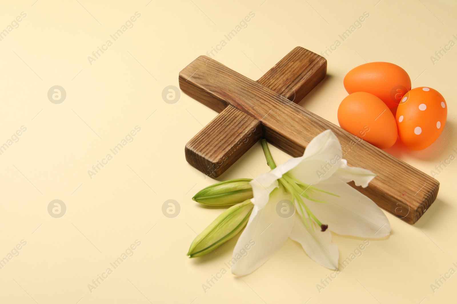 Photo of Wooden cross, painted Easter eggs and lily flowers on pale yellow background, space for text