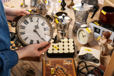 Photo of Woman holding vintage alarm clock near table with different stuff, closeup. Garage sale