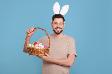 Photo of Happy man in bunny ears headband holding wicker basket with painted Easter eggs on turquoise background
