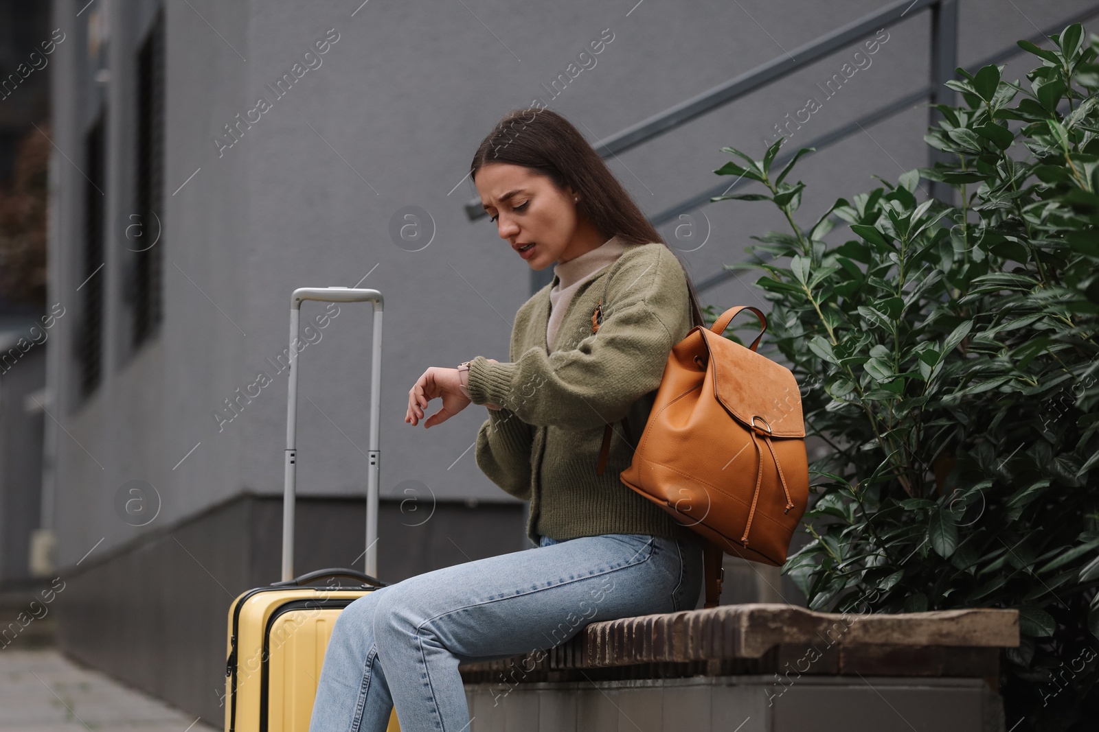 Photo of Being late. Worried woman with suitcase looking at watch on bench outdoors