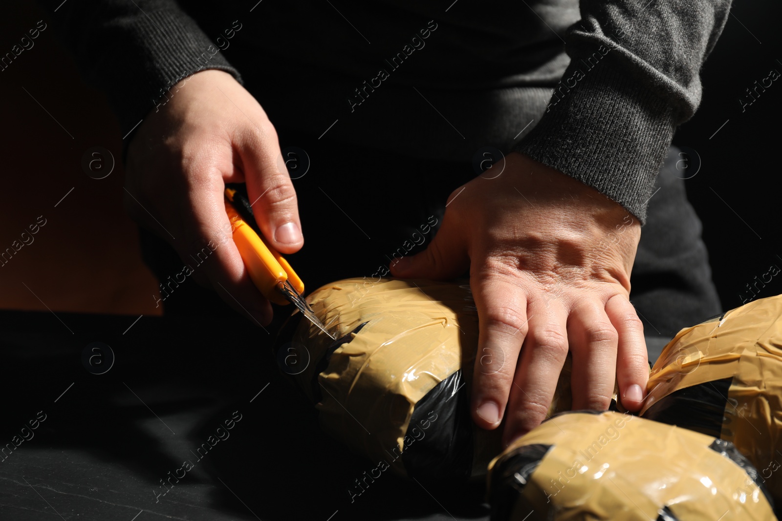 Photo of Smuggling and drug trafficking. Man opening package of narcotics with box cutter in darkness, closeup