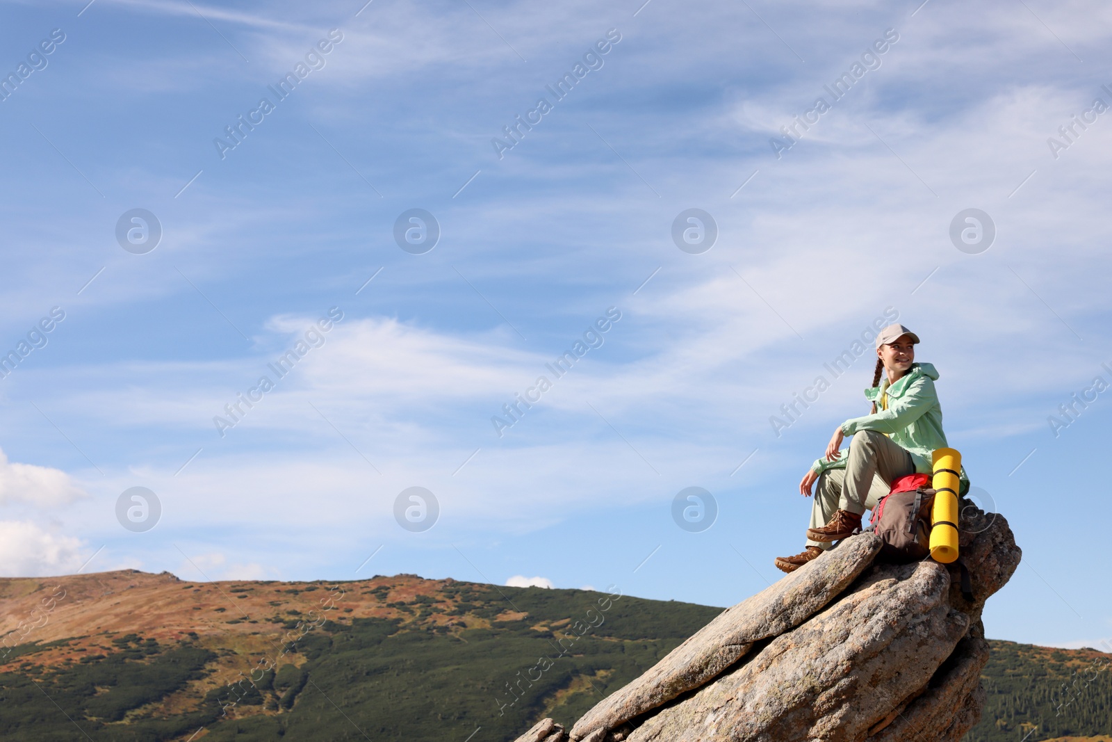 Photo of Young woman with backpack and sleeping mat on cliff in mountains. Space for text