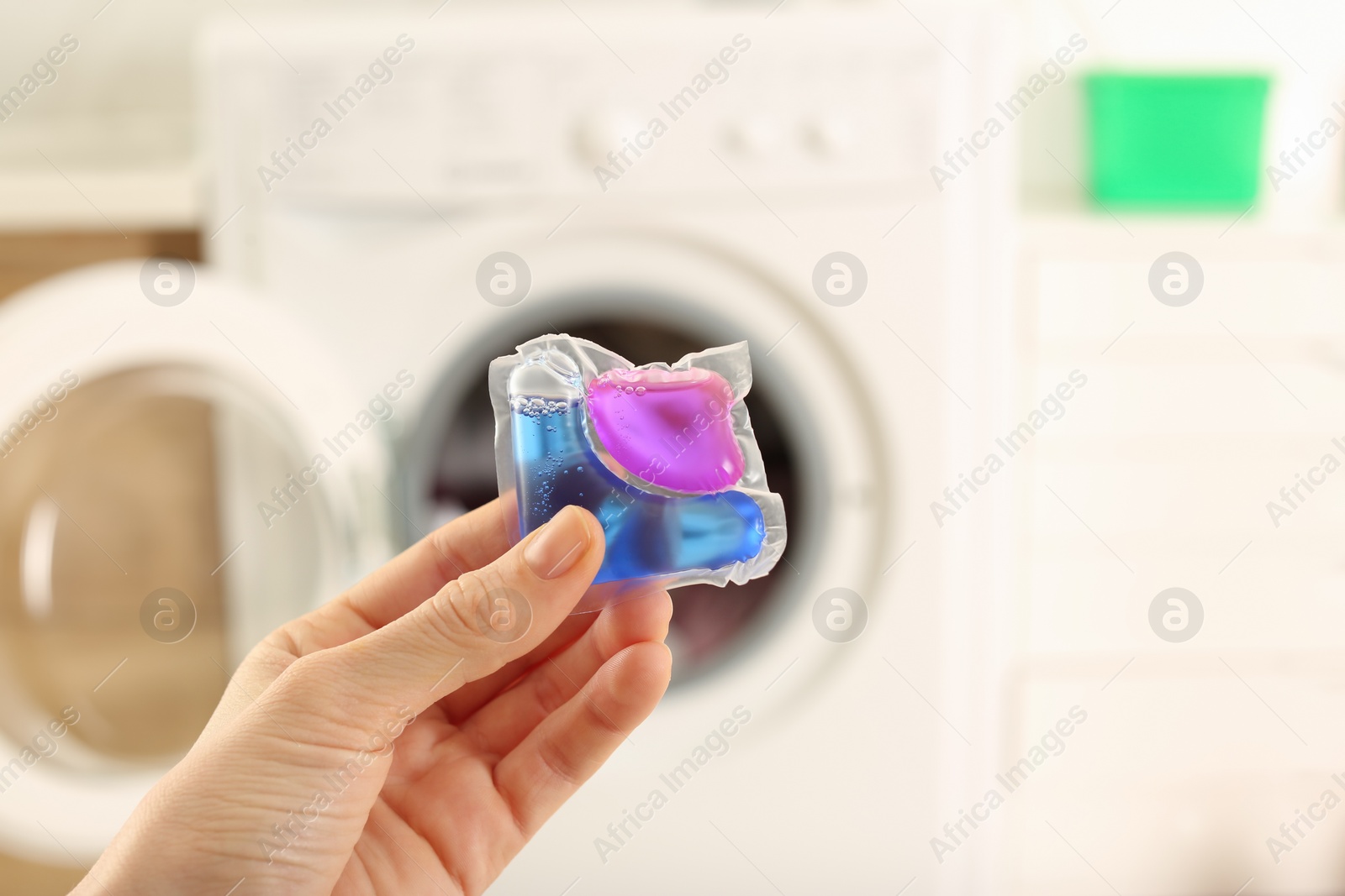 Photo of Woman holding laundry detergent capsule near washing machine indoors, closeup