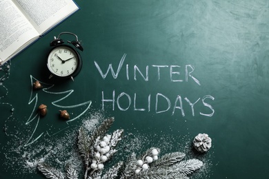 Photo of Flat lay composition of Christmas decorations, book and alarm clock on school blackboard with phrase Winter Holidays