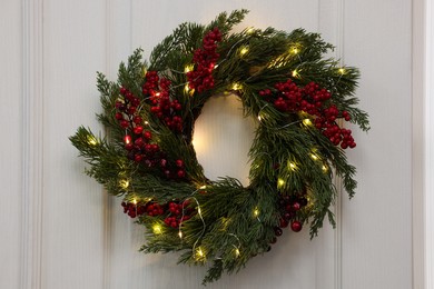 Beautiful Christmas wreath with red berries and fairy lights hanging on white door