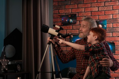 Little boy with his grandfather using telescope to look at stars in room