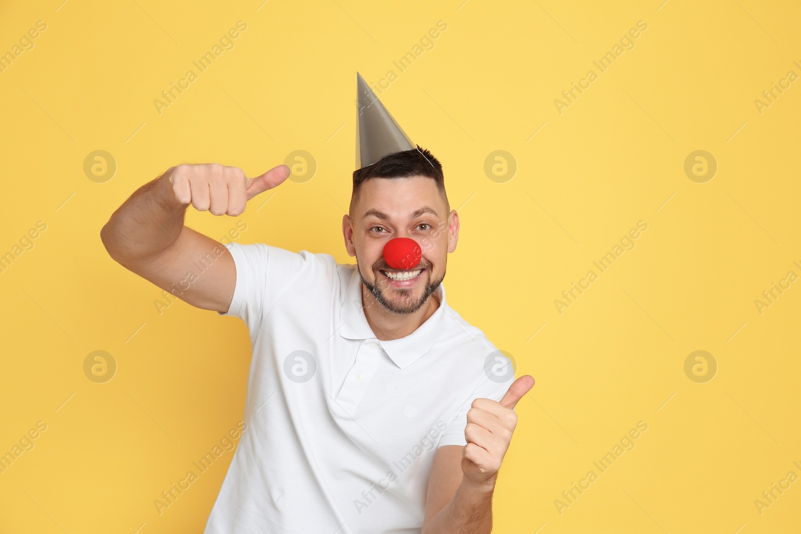 Photo of Funny man with clown nose and party hat on yellow background. April fool's day