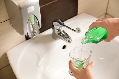 Photo of Man pouring mouthwash from bottle into glass in bathroom, closeup. Teeth care