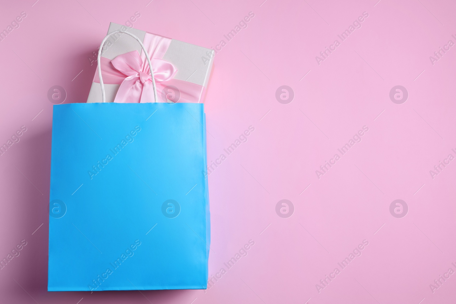 Photo of Light blue paper shopping bag with gift box on pink background, top view. Space for text