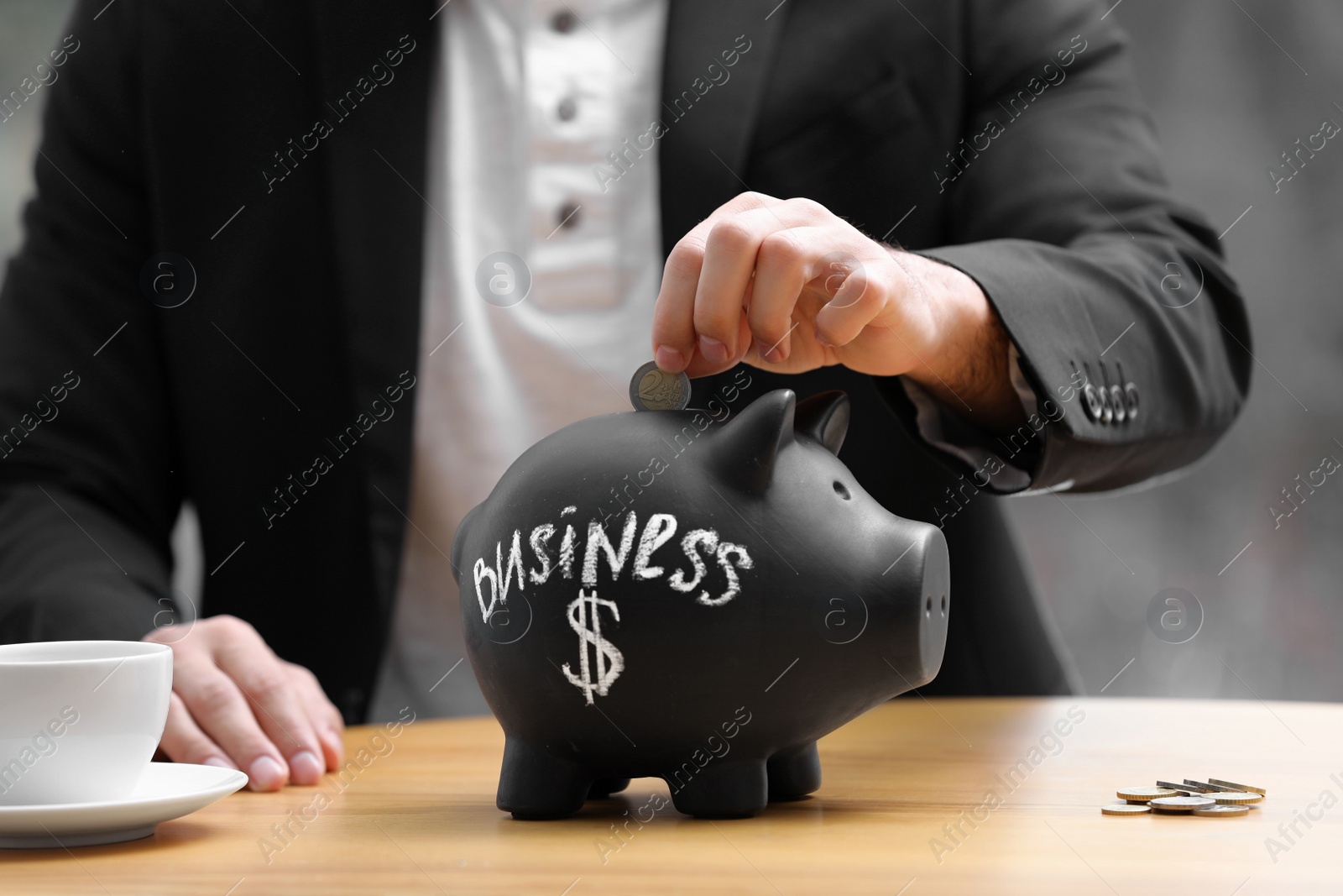 Photo of Man putting money into piggy bank with word BUSINESS at table, closeup