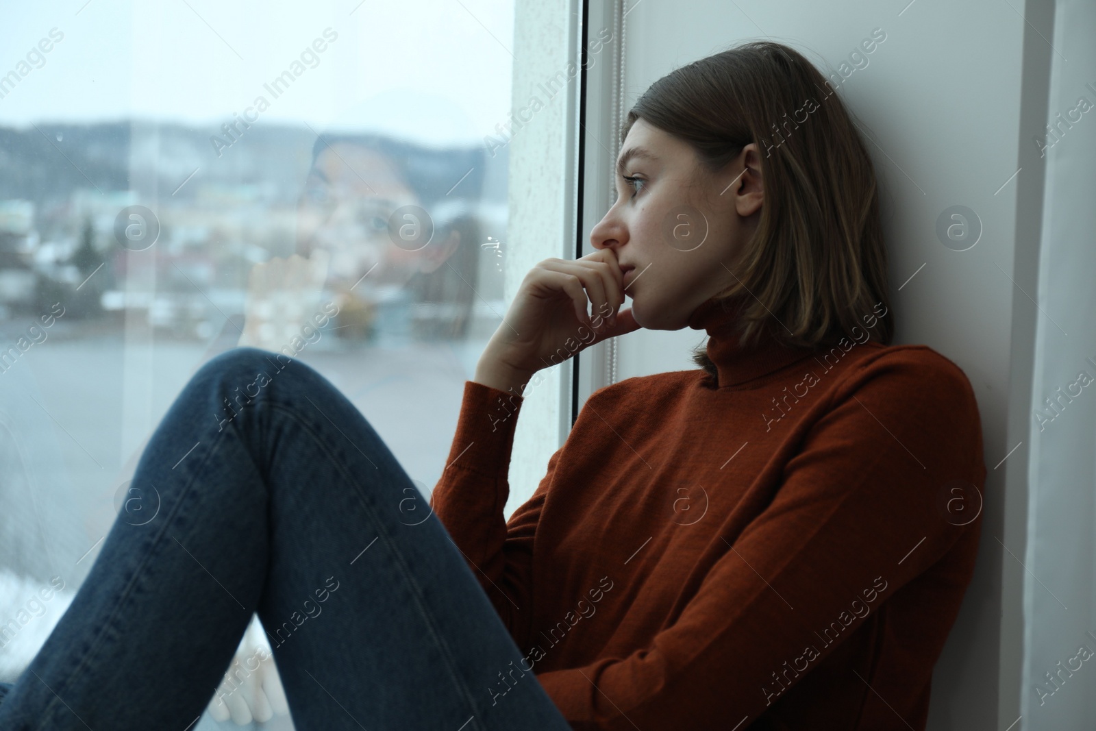 Photo of Sad young woman sitting on windowsill near window at home