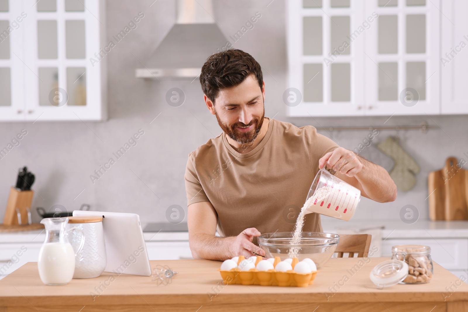 Photo of Man making dough while watching online cooking course via tablet in kitchen