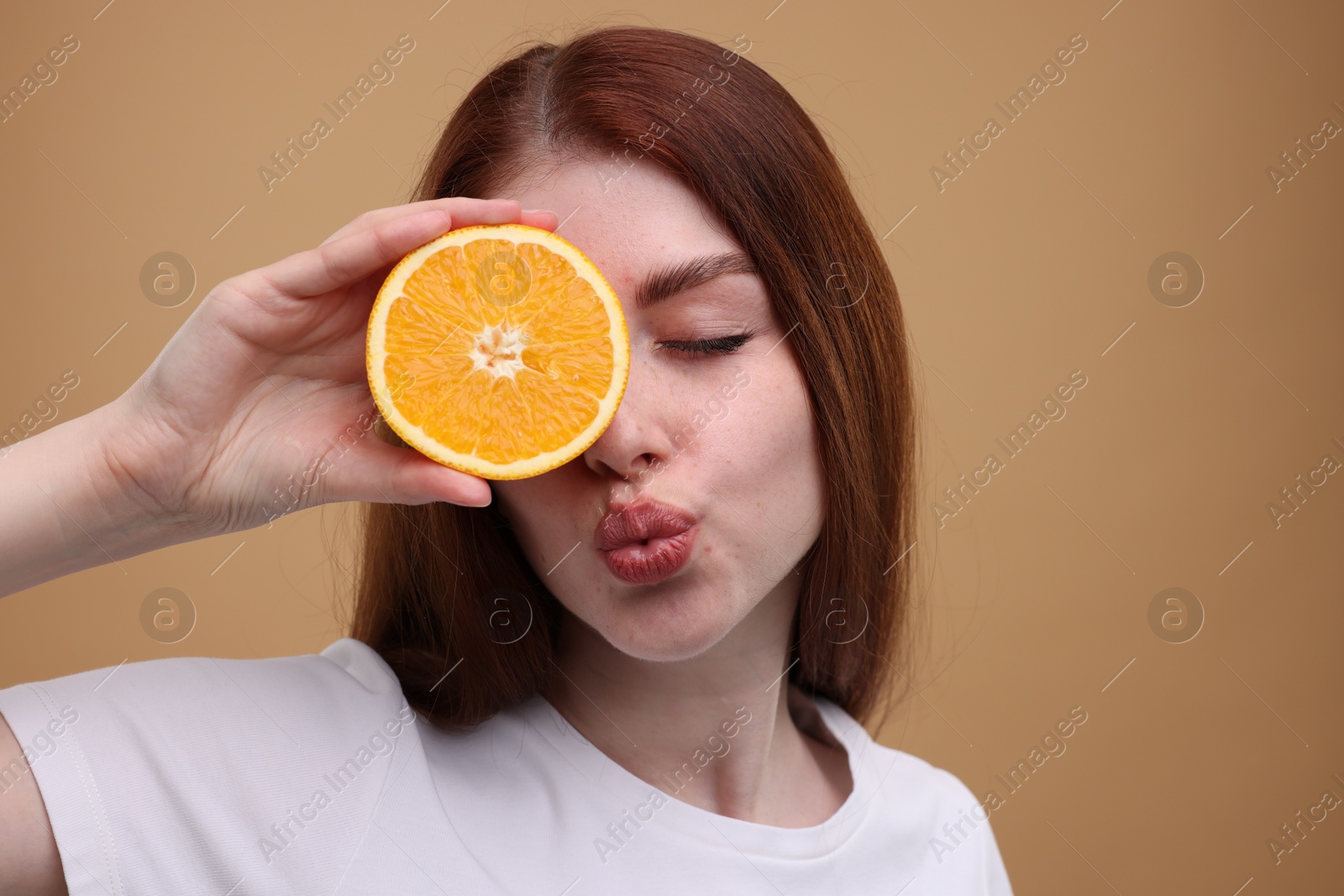 Photo of Beautiful woman covering eye with half of orange and sending air kiss on beige background