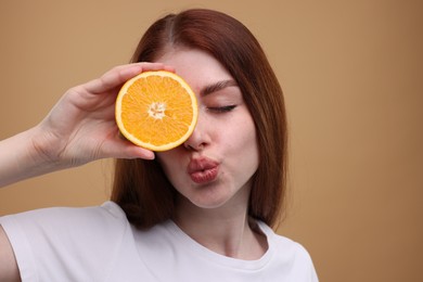 Beautiful woman covering eye with half of orange and sending air kiss on beige background