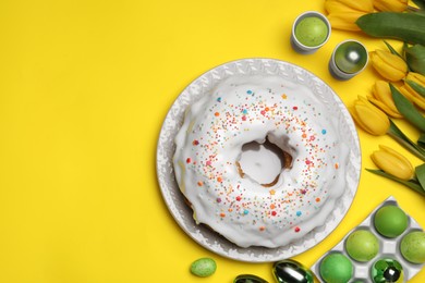 Photo of Easter cake with sprinkles, painted eggs and tulips on yellow background, flat lay. Space for text