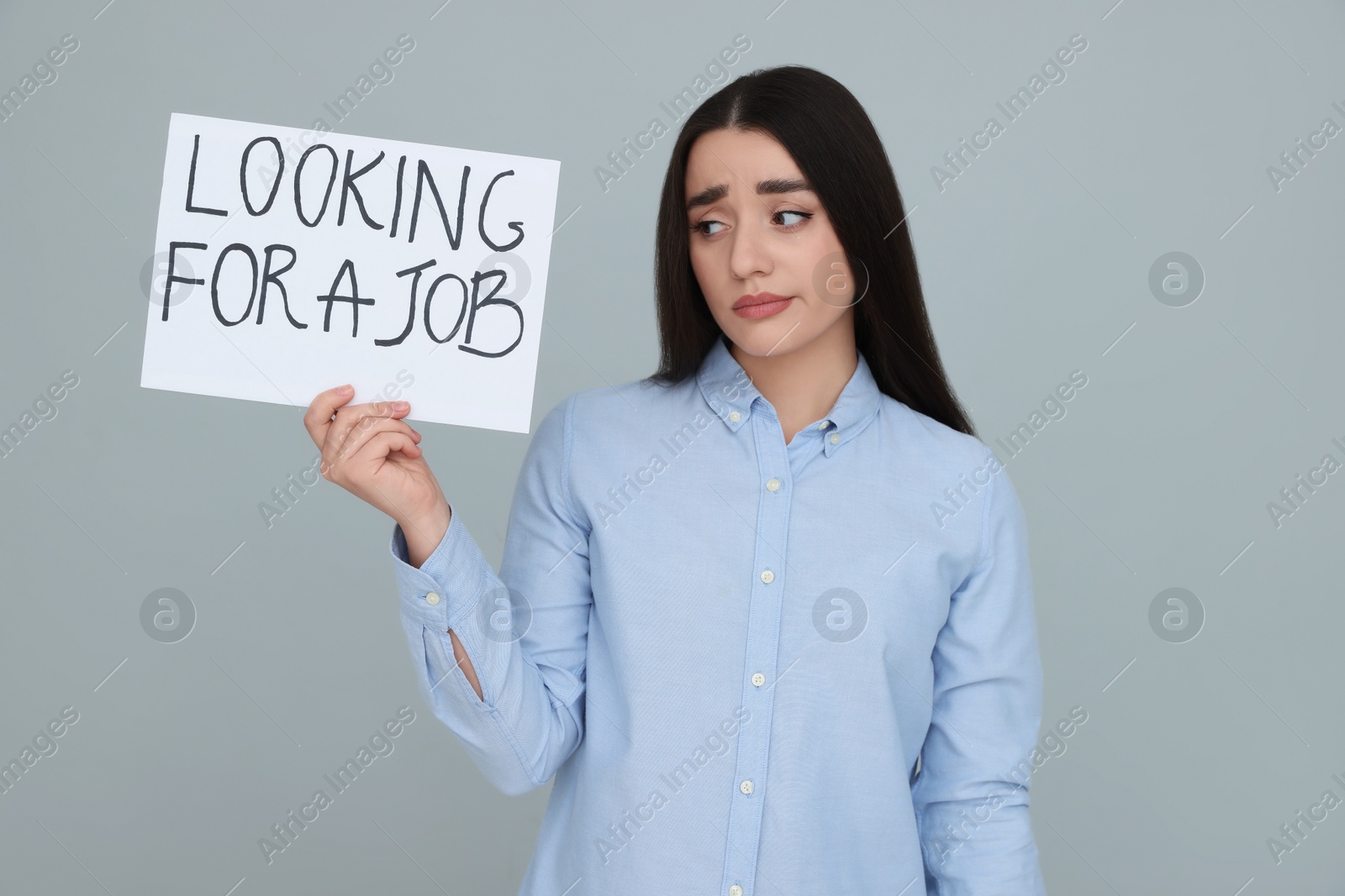 Photo of Young unemployed woman holding sign with phrase Looking For A Job on grey background