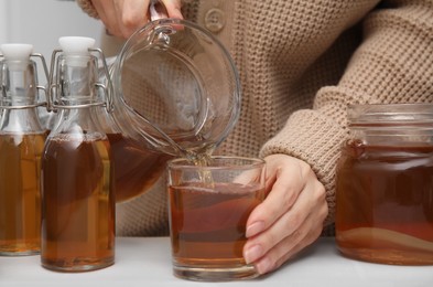 Photo of Woman pouring fermented kombucha from jug into glass at white table, closeup