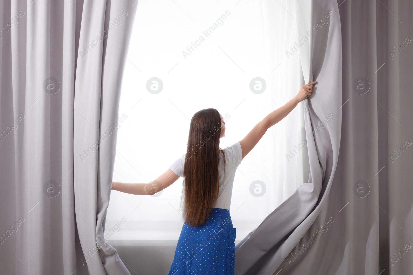 Photo of Woman opening stylish curtains at home, back view