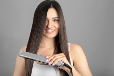 Young woman using hair iron on grey background, space for text