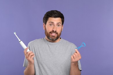 Photo of Confused man with tongue cleaner and electric toothbrush on violet background