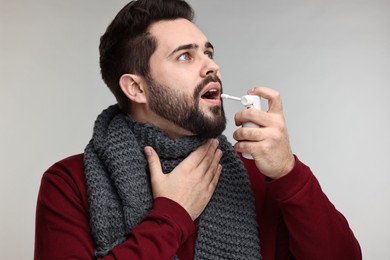 Young man with scarf using throat spray on grey background
