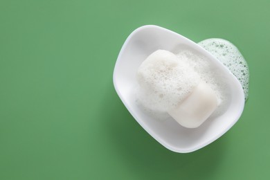 Photo of Dish with soap bar and fluffy foam on green background, top view. Space for text