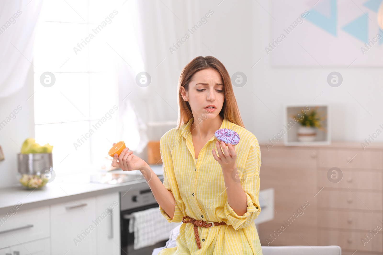 Photo of Thoughtful young woman choosing between orange and donut in kitchen. Healthy diet