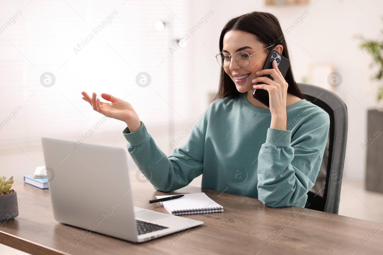 Photo of Young woman talking on smartphone during webinar at table in room