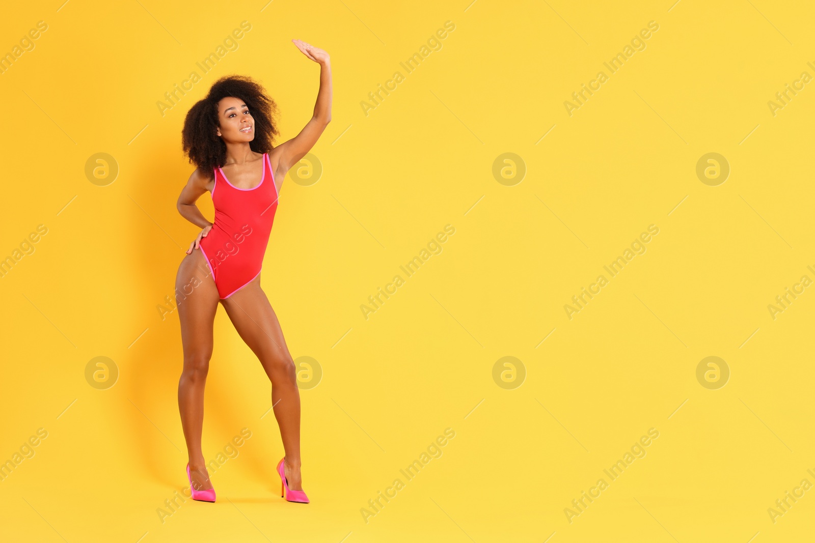 Photo of Beautiful woman in bright one-piece summer swimsuit and stylish high heel shoes on yellow background, space for text