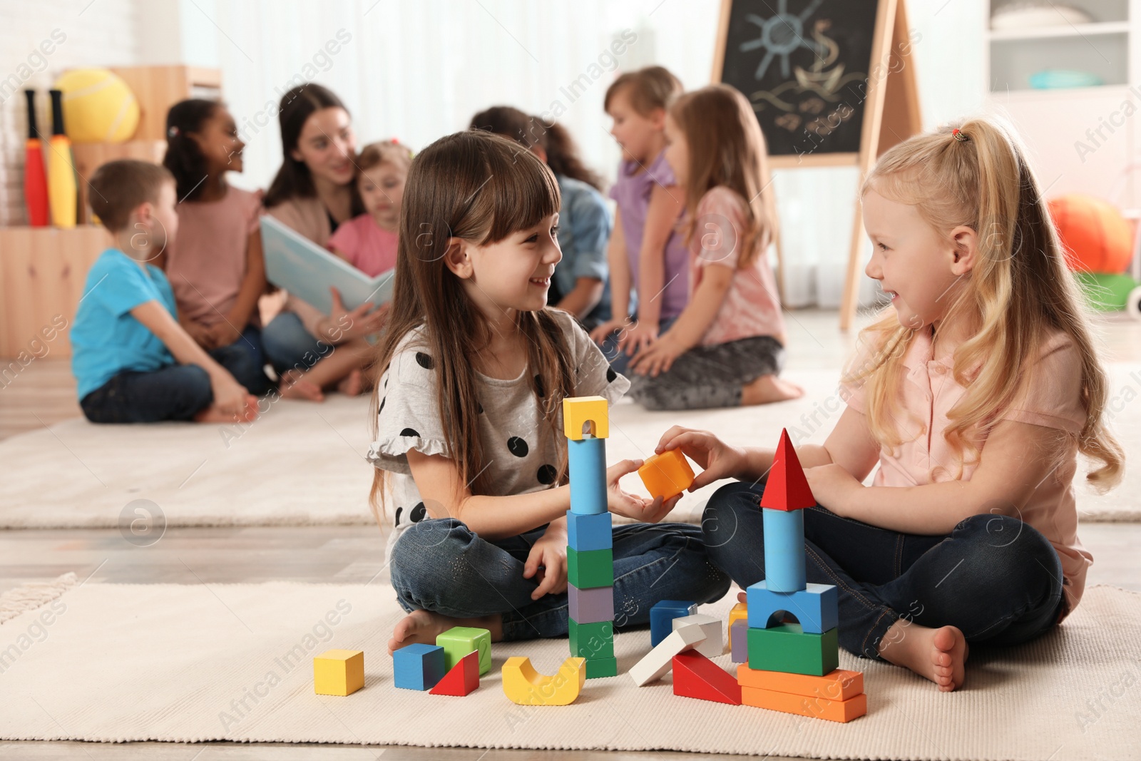 Photo of Cute girls playing with building blocks on floor while kindergarten teacher reading book to other children indoors