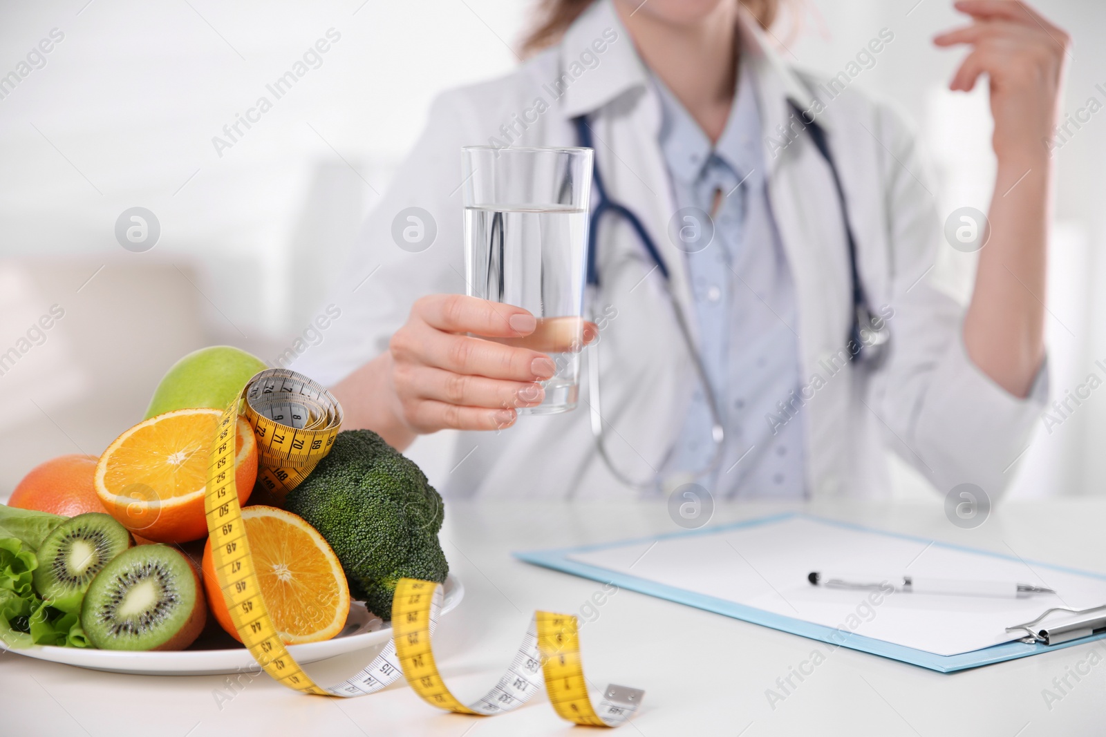 Photo of Nutritionist with glass of water, fruits, vegetables and measuring tape in office, closeup