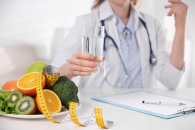 Nutritionist with glass of water, fruits, vegetables and measuring tape in office, closeup