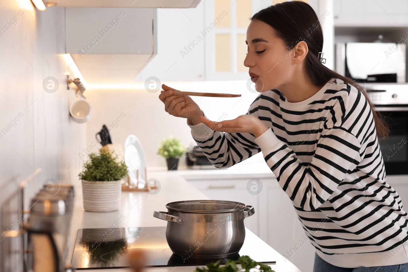 Photo of Beautiful woman with wooden spoon tasting soup in kitchen