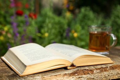 Photo of Open book with glass cup of tea on wooden table in garden