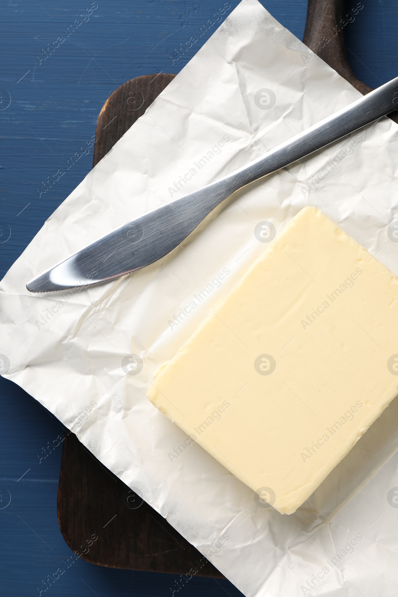 Photo of Block of tasty butter in open foil packaging and knife on blue wooden table, top view