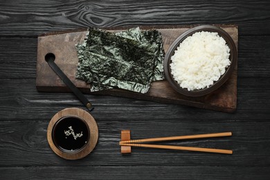 Photo of Dry nori sheets, rice, soy sauce and chopsticks on black wooden table, flat lay