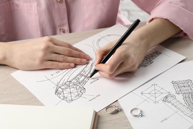 Jeweler drawing sketch of elegant ring on paper at wooden table, closeup