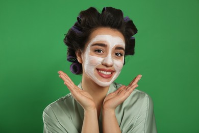 Photo of Happy young woman in silk bathrobe with hair curlers and facial mask on green background