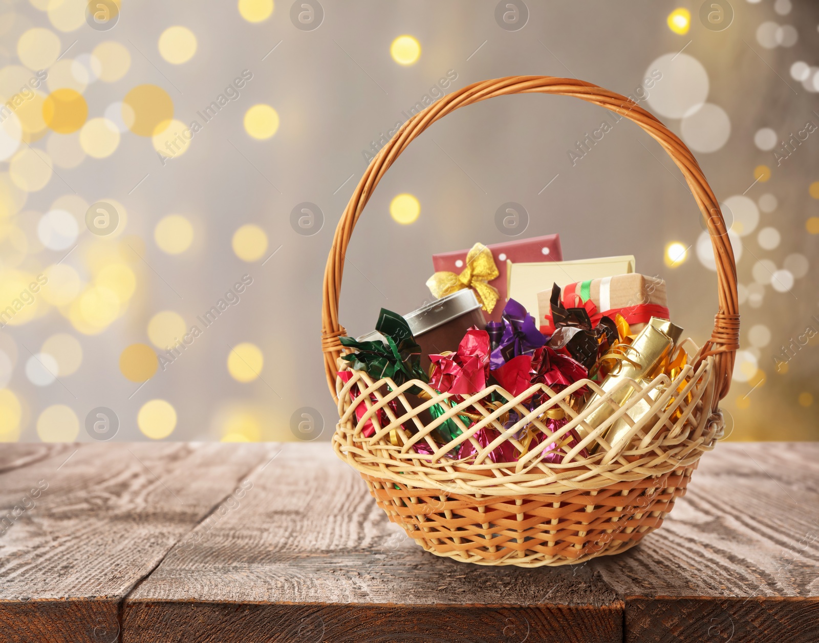 Image of Wicker basket full of gifts on wooden table against blurred festive lights. Space for text