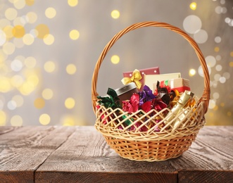 Image of Wicker basket full of gifts on wooden table against blurred festive lights. Space for text