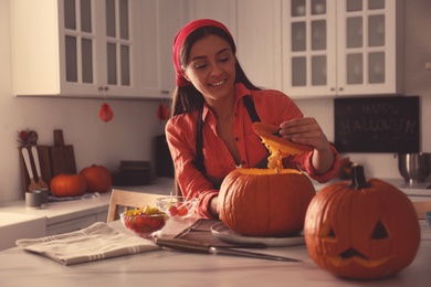 Photo of Woman making pumpkin jack o'lantern at table in kitchen. Halloween celebration