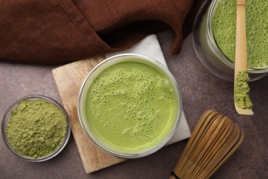 Photo of Tasty matcha smoothie, powder, bamboo spoon and whisk on brown table, flat lay